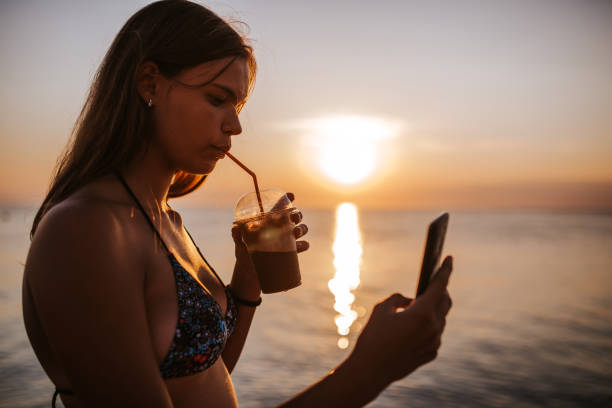 istock Teenage girl drinking coffee and using mobile phone on beach during beautiful sunset 1160261949