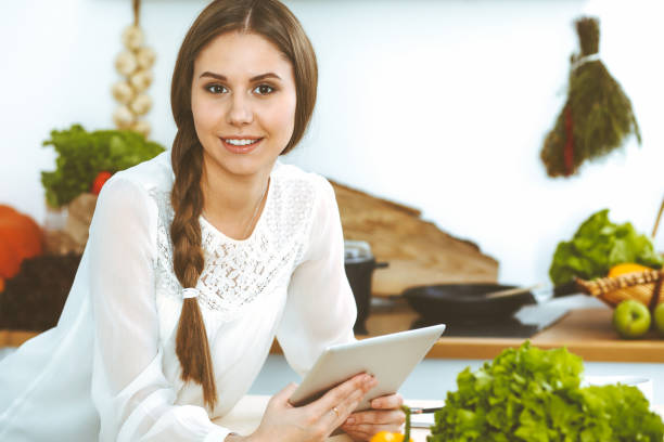 istock Young woman using tablet while cooking or making online shopping in kitchen. Girl looking at the camera. Healthy lifestyle and vegan meal concepts 1160331930