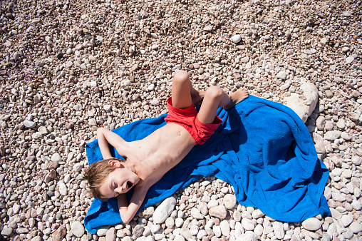 happy funny little kid in red shorts lying on blue towel on pebble summer sunny beach yawning