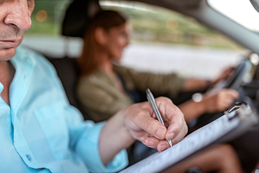 Close up of Driving instructor and woman student in examination car. Learner driver student driving car with the instructor. Driving test. Young smiling woman driving car feeling inexperienced, looking nervous at the road traffic for information to make appropriate decisions. Senior Man is an instructor, controlling and checking while holding a clipboard.