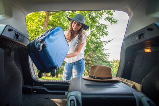 Girl packing car for road trip. She put suitcase in the back of the car. Girl have curly hair and hat on the head. Natural ambient and modern car with girl in casual outfit. This photo was captured on the bright and warm day.