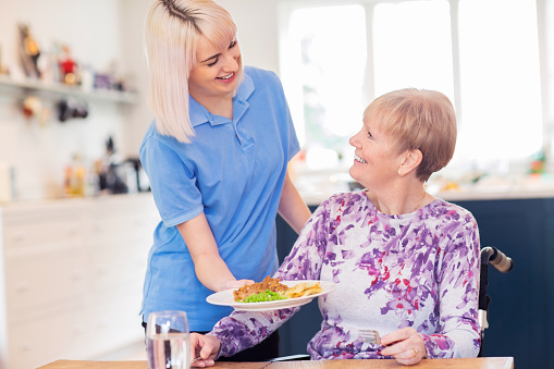 Asistente de cuidado femenino sirviendo comida a una mujer mayor sentada en silla de ruedas en la mesa photo