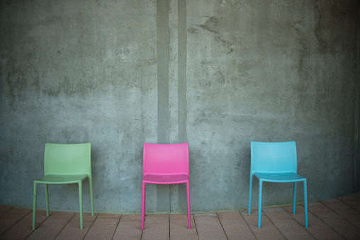 Three multi colored chairs outdoors, against a concrete wall, waiting for someone to sit on them