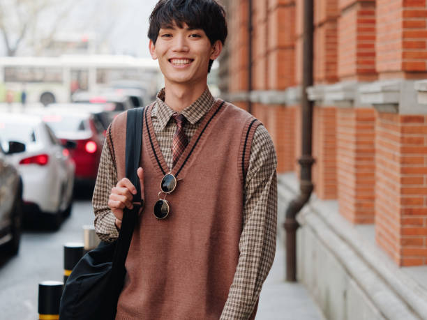 Portrait of a handsome Chinese young man with Korean style clothes smiling and looking at camera confidently with street background, male fashion, cool Asian young man lifestyle stock photo