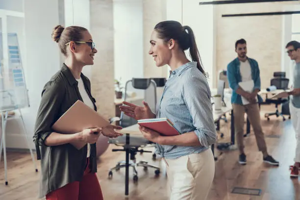 Waist up portrait of smiling female colleagues talking in meeting room and holding laptop and notebook