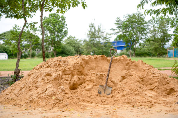 A sizable mound of dirt bordered by a black plastic barrier sits at a construction site, with mature trees and a quiet suburban house in the background, bathed in warm evening light.