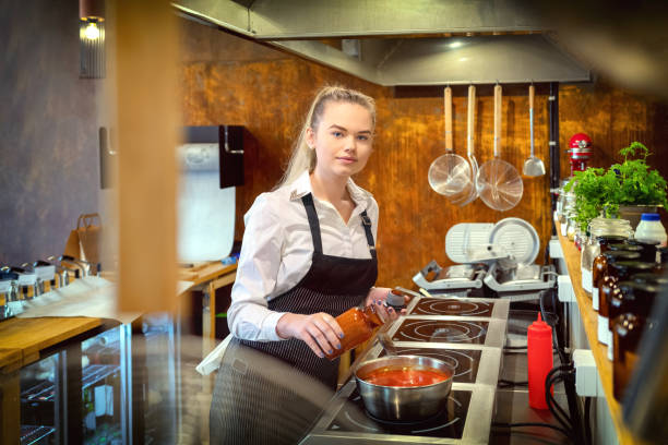 istock Happy satisfied woman adding condiments for taste in boiling pot with pasta sauce - small business woman looking at camera and cooking in restaurant kitchen - chef in apron preparing food recipe 1160420902