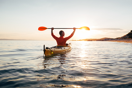 Rear view of senior man in kayak holds paddles high.