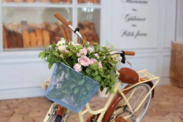 old bicycle with a basket of roses against the wall in pastel colors. Decorative bicycle stand for plants and flowers. Beautiful romantic landscape: vintage wicker basket with flowers near the café.