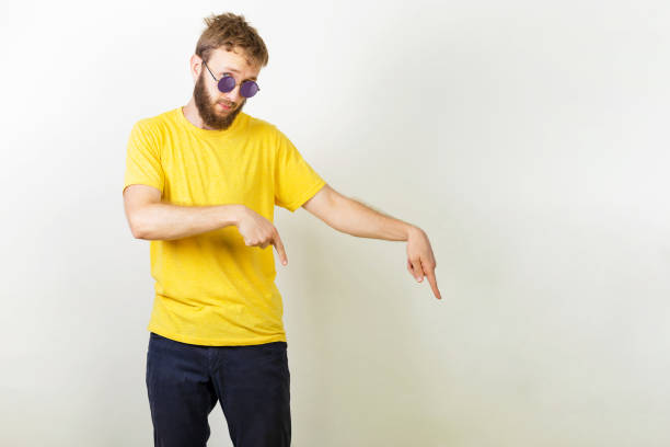 istock Bearded man in a yellow T-shirt and round sunglasses showing direction and points down with both fingers, advertises new material for floor, People, lifestyle, promotion concept 1160438310