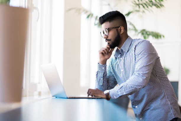istock Indian young man with laptop standing in modern office background 1160438389