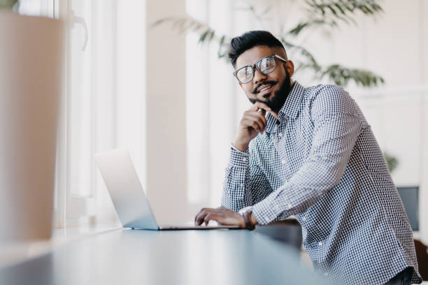 istock Indian young man with laptop standing in modern office background 1160439120