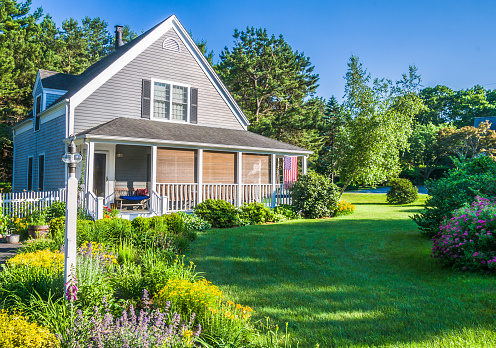 A colorful summer flower garden and landscaping enhance the setting of this Cape Cod home which proudly displays the American Flag and has rattan shades on the open front porch.