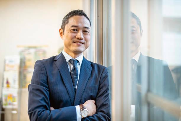 Male Japanese Hospital Administrator Standing at Window Smiling male hospital administrator in a blue suit taking a break from his busy day to look out an office window. east asia photos stock pictures, royalty-free photos & images