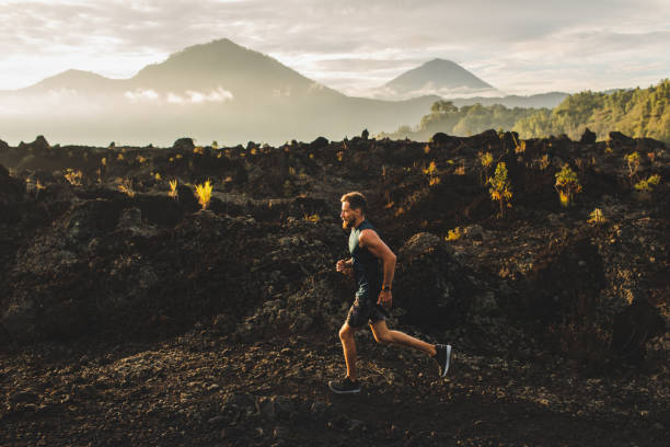 日の出時に山の中を走る若い男性アスリートトレイル。背景にバリ島の驚くべき黒い溶岩火山の風景。アドベンチャースポーツコンセプト。 - footpath single lane road sunrise landscape ストックフォトと画像