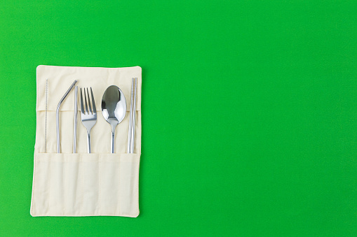 Reusable and eco-friendly silver cutlery set; Chopsticks, spoon, fork, straw and tube brush in separated sockets pouch. Flat lay shot on a green background with copy space.