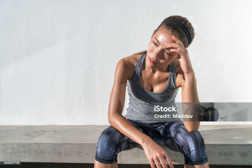 Tired fitness running woman sweating exhausted Tired fitness running woman sweating taking a break of cardio workout difficult training. Exhausted runner sitting with headache feeling exhaustion and dehydration from working out at outdoor gym. Exercising Stock Photo