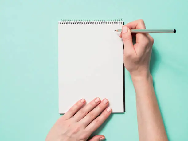 Blank paper notebook and female hands with pencil. Empty paper sketchbook with pincil in woman hands on blue background. Top view or flat lay. Copy space for text or design.