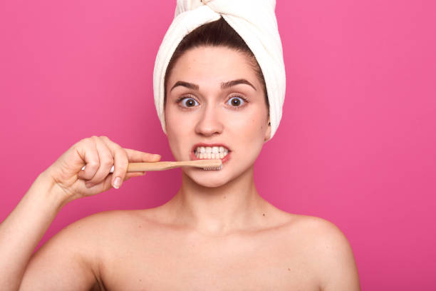 istock Attractive woman brushing her teeth with toothbrush and toothpaste in bathroom, doing morning procedures with bare shoulders and wrapped towel, has astonished facial expression, isolated over pink. 1160555357