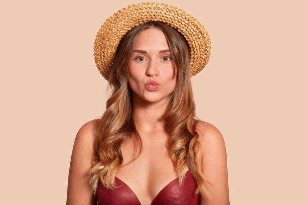 Beautiful woman wearing summer straw hat, makes kissing gesture, keeps lips rounded, attractive female with perfect tanned skin, posing isolated over beige studio background with copys pace.