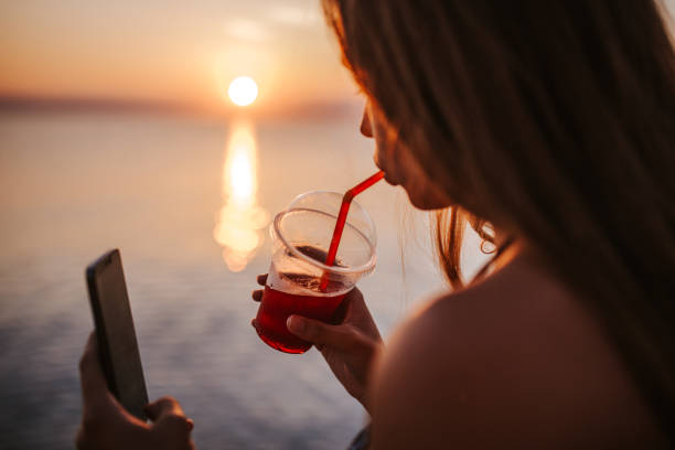 istock Teenage girl drinking coffee and using mobile phone on beach during beautiful sunset 1160557578