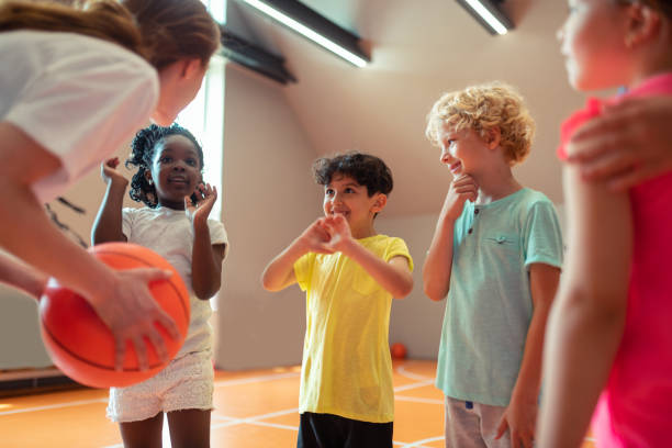 Children smiling to their sports teacher in the gym. Best teacher. School children smiling to their sports teacher playing basketball in the gym. youth basketball indoor stock pictures, royalty-free photos & images