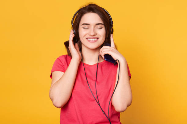 istock Smiling cheerful young female standing isolated over yellow background in studio, closing her eyes while listening to music, having headphones, getting pleasure, wearing casual red t shirt. Free time. 1160563996