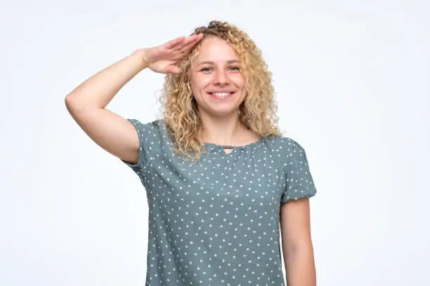 Yes sir. Young worker woman with curly hairstyle looking at camera with salute and ready to do your order. Positive facial emotion.