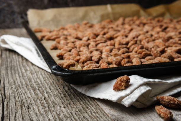 Burnt almonds just taken out of the oven - horizontal Burnt almonds outspread on black baking tray, paper, white cloth on rustic textured wooden table, three burnt almonds laying in foreground, dark background - angled view, landscape orientation candied fruit stock pictures, royalty-free photos & images