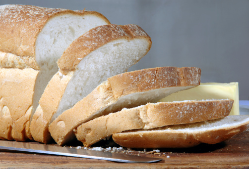 Crusty loaf of finest bread on a bread board with a platter of butter in the background and the knife that sliced it in the foreground. Good sharpness through the image.