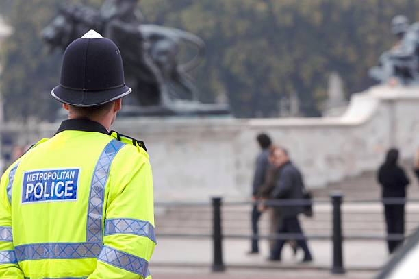 A British cop standing guard at Buckingham Palace A British police man stands on duty across the road from Buckingham Palace, as tourists pass by in the background on their way to see the Changing of the Guard. metropolitan police stock pictures, royalty-free photos & images