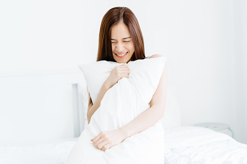 Portrait of smiling beautiful young Asian woman holding a pillow and close your eye on bed. Good morning, new day, weekend, holidays concept.