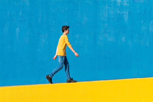 Young boy wearing casual clothes walking against a blue wall in a sunny day