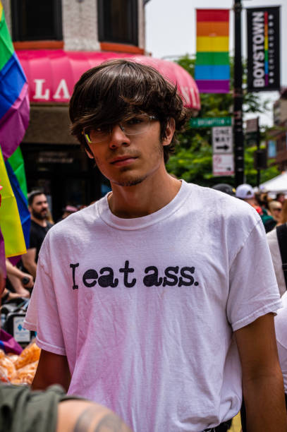 A confident young man is wearing a white tee shirt that reads "I eat ass." at the Gay Pride parade. stock photo