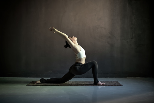Woman doing hatha yoga asana Anjaneyasana or low crescent lunge pose. Studio shot on black wall full length