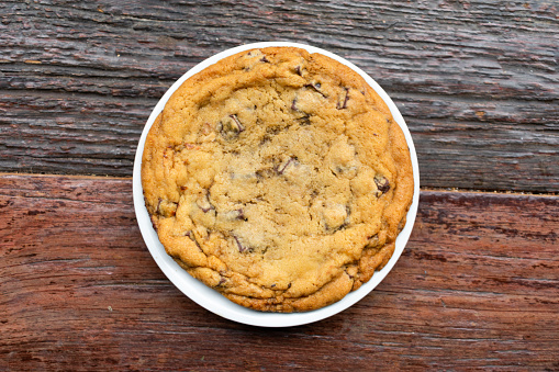 A large chocolate chip cookie on a white plate on top of a rustic wooden table