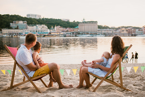 Family at seaside in evening open-air cafe. Mother and father and two sons sit on sun loungers, looking at sunset on sandy beach near river overlooking city. Concept travel and summer family vacation.