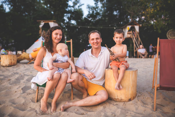 Young beautiful Caucasian happy family of four sitting together embracing at beach cafe in evening sunset. Theme family vacation and family values. Two brother boys and mom, dad sit on sandy beach. joven hermosa familia caucásica feliz de cuatro sentados juntos abrazando en la cafetería de la playa en la puesta de sol de la noche. temática vacaciones familiares y valores familiares. dos hermanos y mamá, papá se sente en la playa de arena