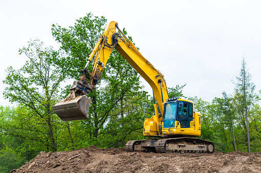 Hydraulic earth mover with rotating cabin, shovel and continuous tracks. Pile of soil, spring sky, green tree tops. Building industry