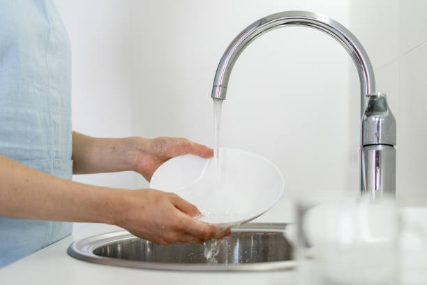 Cropped view of woman holding dishware in hands, washing plate under water in sink, standing on white bright kitchen
