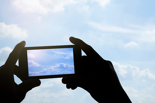 Silhouette of hands holding smart phone photographing blue sky with clouds