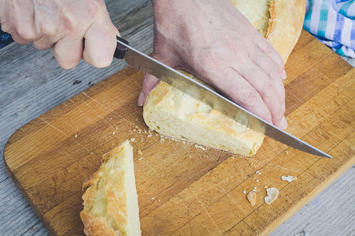 Senior woman cutting freshly baked bread on the wooden board on the rural kitchen table. Traditional bakery concept. Rustic vintage style.