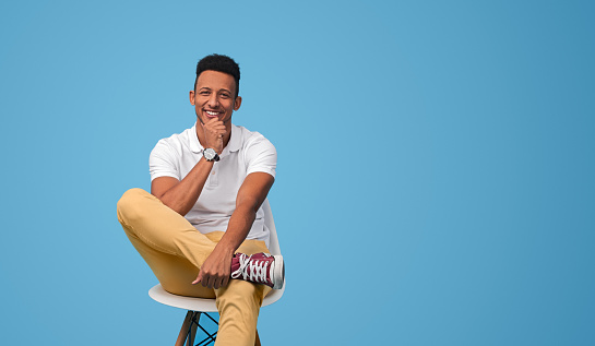 Confident African male in casual outfit rubbing chin and smiling while sitting on chair near empty space on blue background