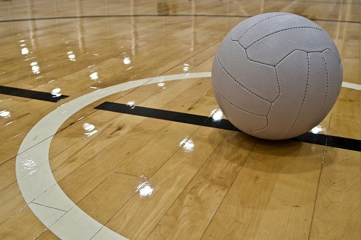 Placed within the white centre circle line markings of an indoor Netball Court, a white Netball Ball is ready to commence the next competition game. The glossy, varnished hardwood floor, reflects the indoor centre's ceiling lights.