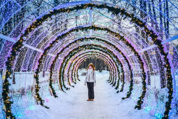 Photo of A pretty woman is standing on the path under the Christmas lights in the winter park.