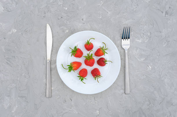 Red ripe strawberries berry on white plate and cutlery, fork, knife. Still life on gray stone background Top view, flat lay, copy space. Concept sweet summer menu, detox, diet