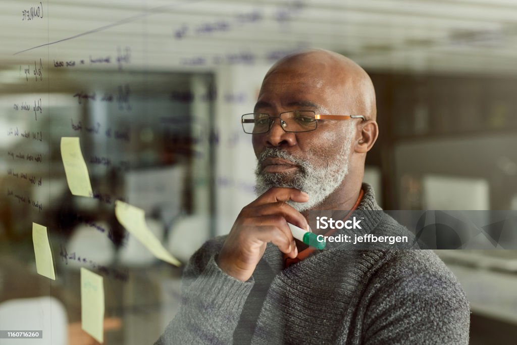 With my expertise, I'll have this sorted in no time Cropped shot of a mature businessman brainstorming with notes on a glass wall Contemplation Stock Photo