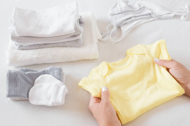 白いベッドシーツの上に新しい赤ちゃんの服を選別する女性の手。母の日常業務柔らかい色。クローズ アップ。 - onesie ストックフォトと画像