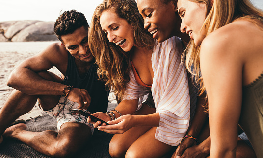 Smiling woman looking at her mobile phone sitting on beach with her friends. Man and three women on a vacation having fun sitting on beach looking at a mobile phone.