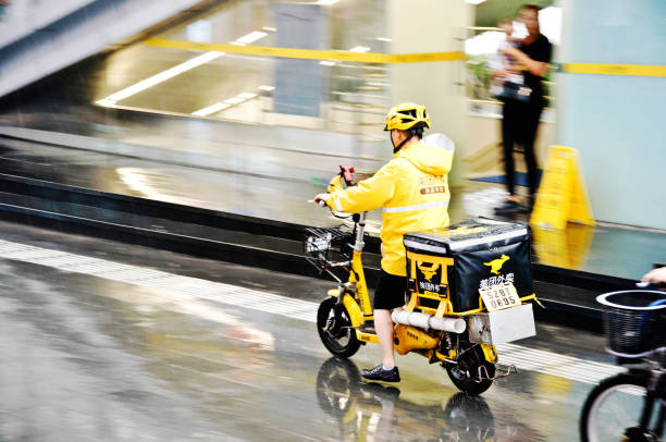 Meituan food delivery worker on motorcycle in the rain in Futian Business District, Shenzhen - China.  Founded by Wang Xing, the app is used in China for food delivery, restaurant deals, movie tickets, hotel, travel bookings, etc A Meituan food delivery worker on motorcycle in the rain in Futian Central Business District, Shenzhen - China. Meituan, founded in 2010 by Wang Xing, is an app that users in China turn to for food delivery, restaurant deals, movie tickets, hotel and travel bookings, and more. chinese script photos stock pictures, royalty-free photos & images